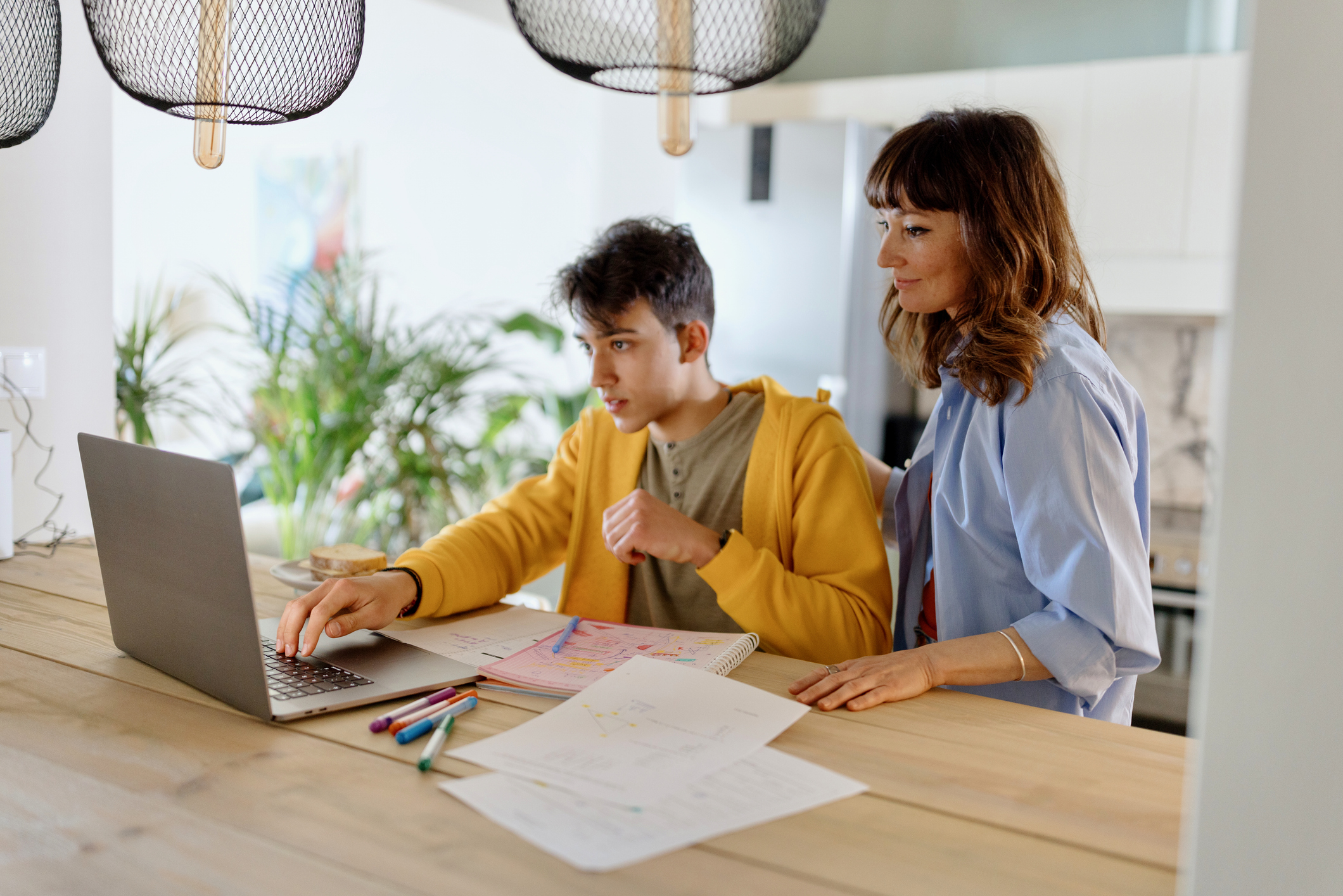 Parent and teenage child look at laptop in kitchen.