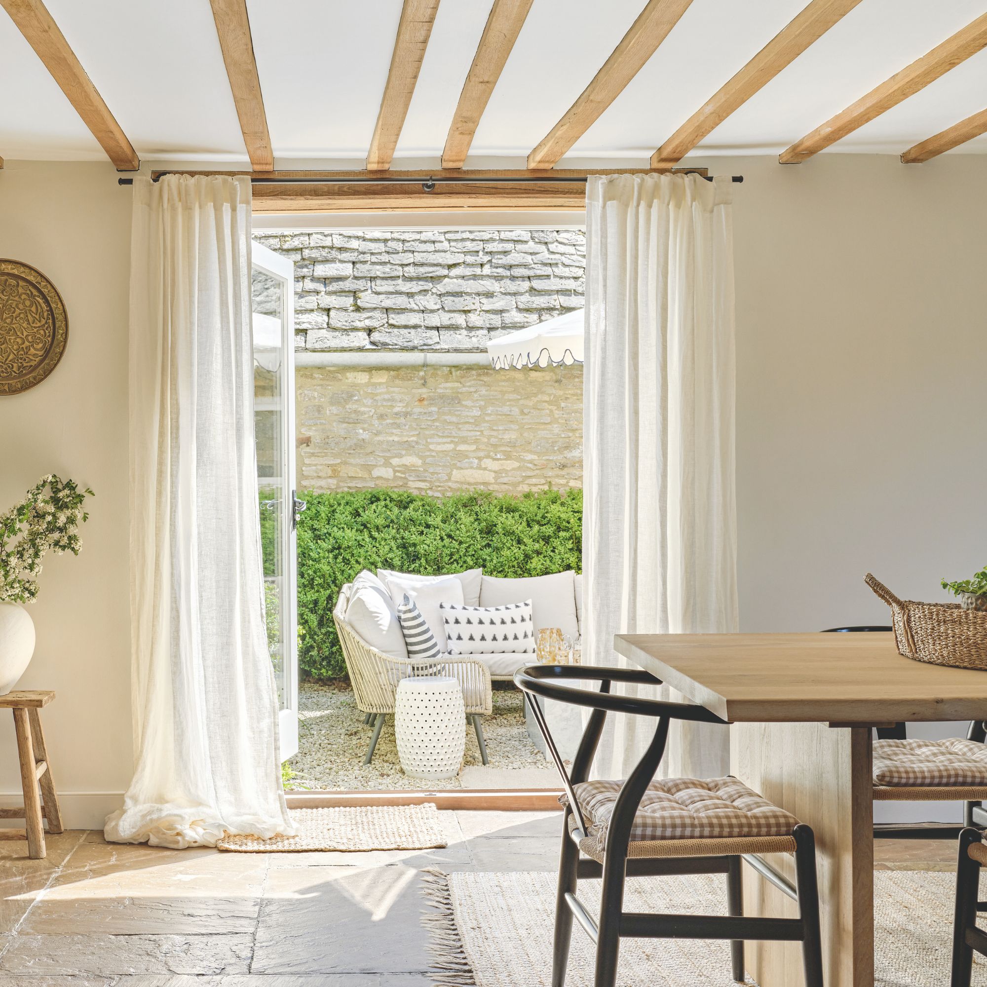 White cottage dining room with wooden beams on ceiling, double doors leading to garden and stone floors with a rug and wooden dining table ontop and black wishbone chairs