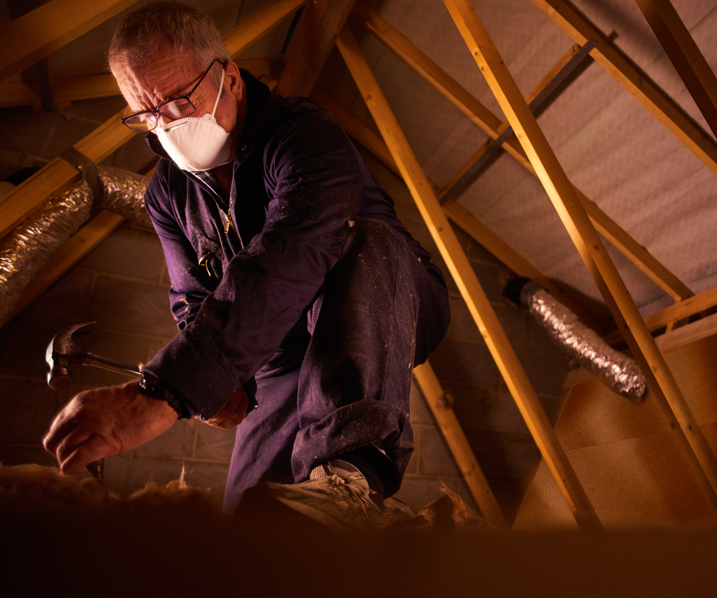 man wearing work overalls and mask with hammer in loft looking at floor