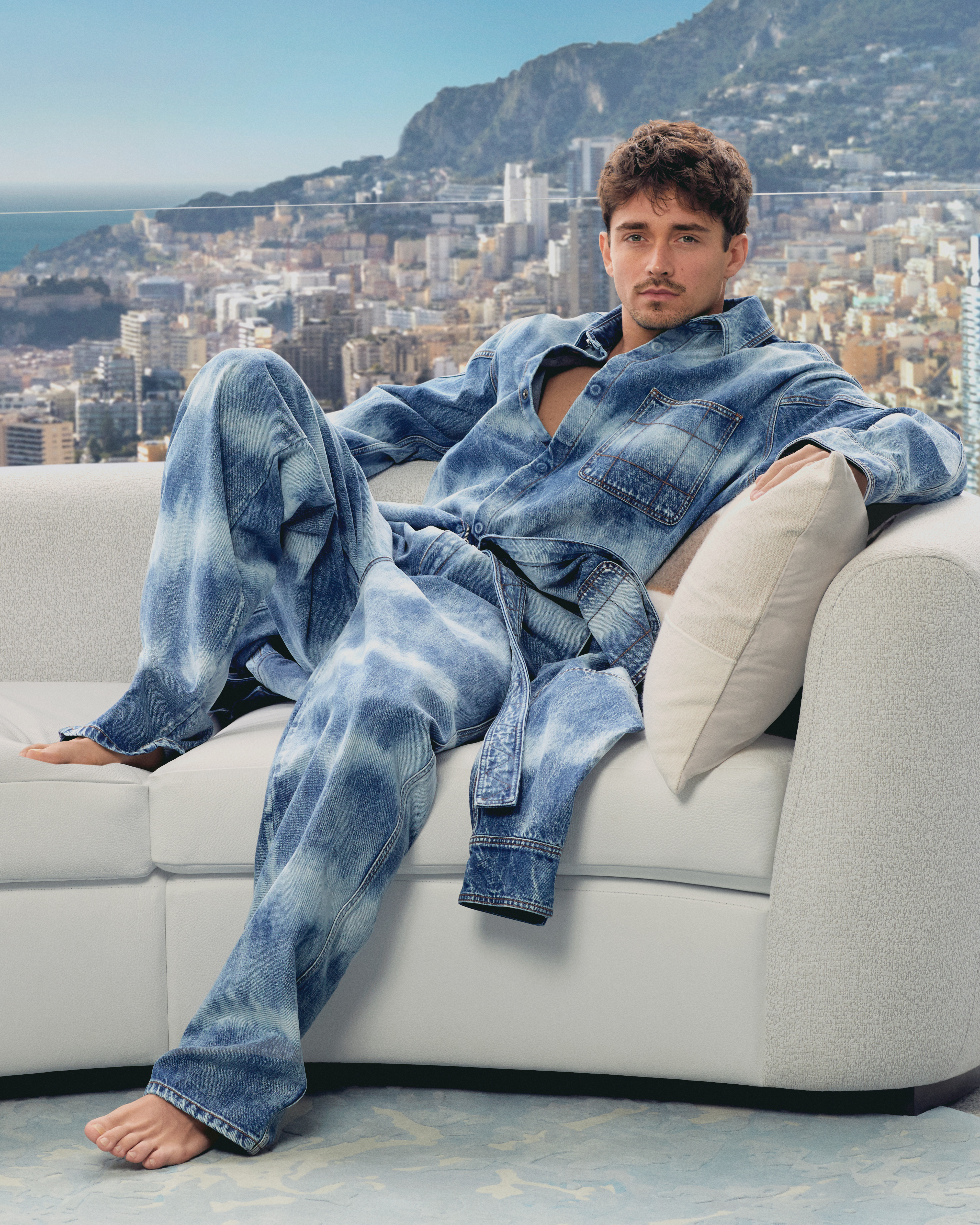 Scuderia Ferrari driver Charles Leclerc posing in his collaboration collection with Ferrari Style on a sofa with Monaco as a backdrop.