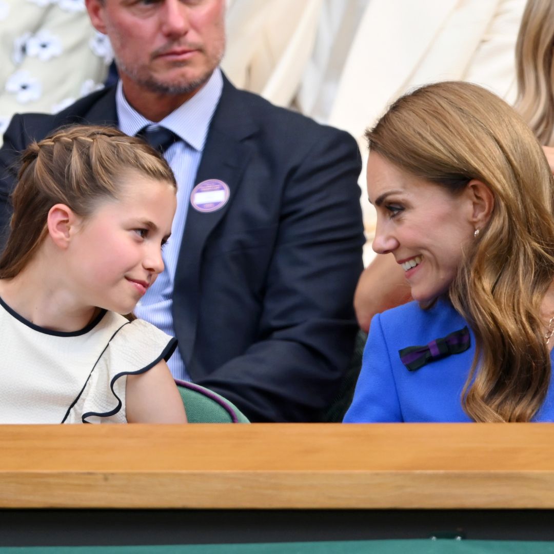 Princess Charlotte wearing a white dress leaning forward and talking to Princess Kate in a blue dress at Wimbledon