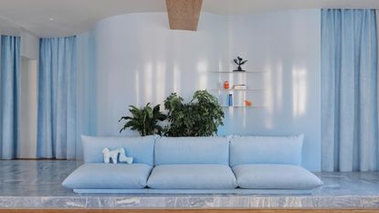 Image of a modern, powder blue living room with glossy walls that have thick velvet curtains over the doorways. There is light blue marble flooring and an overstuffed modern sofa in a light blue velvet. 