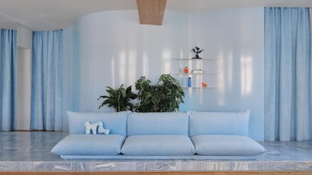 Image of a modern, powder blue living room with glossy walls that have thick velvet curtains over the doorways. There is light blue marble flooring and an overstuffed modern sofa in a light blue velvet.