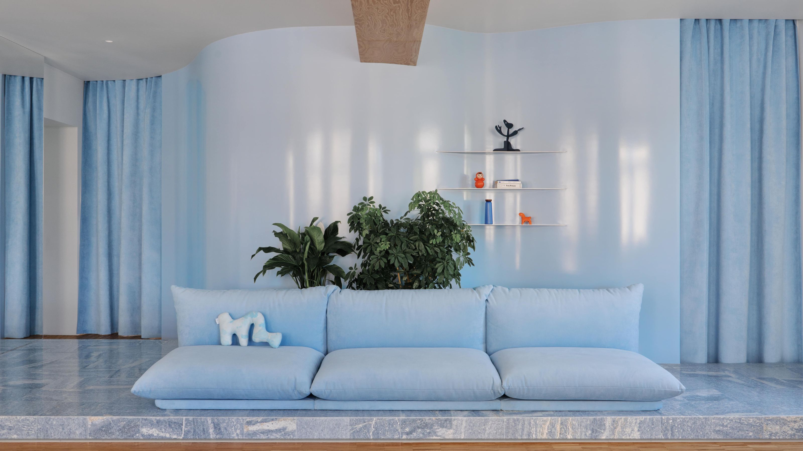 Image of a modern, powder blue living room with glossy walls that have thick velvet curtains over the doorways. There is light blue marble flooring and an overstuffed modern sofa in a light blue velvet. 