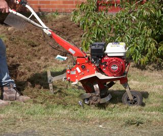 man wearing jeans and safety boots operates petrol driven garden rotavator being used to churn up soil in a garden