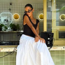Singer Olivia Dean sits on a tabletop wearing a white cotton skirt with Adidas trainers in black and a black sleeveless top.