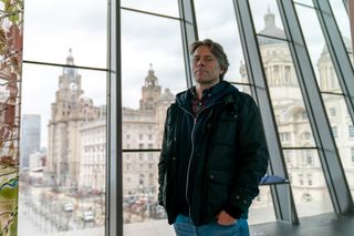 John Bishop as Dan Lewis, standing in front of a wall comprised of glass windows in Liverpool, with the Liver Building visible in the distance behind him