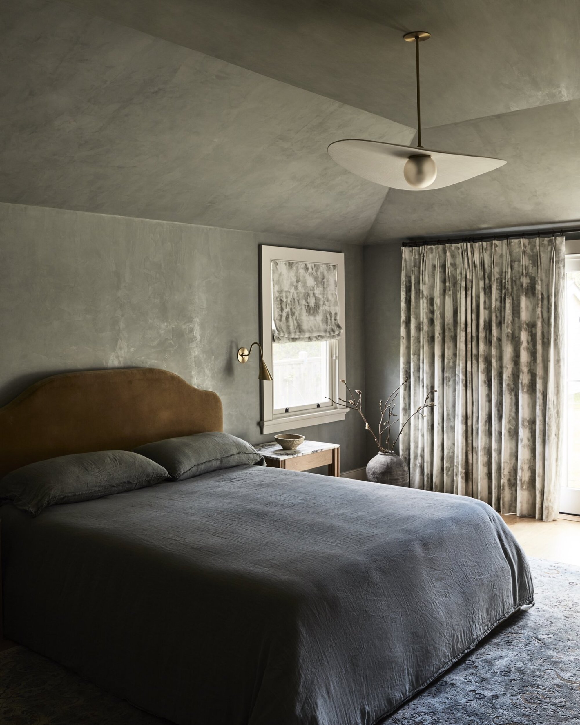 Image of a bedroom painted in a limewash gray with a velvet ochre yellow headboard and bedding that is a textured, wrinkled dark gray.
