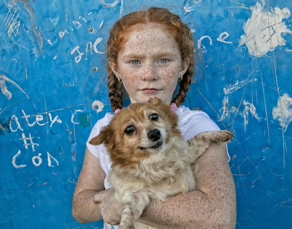 A girl with braided hair holds a small, happy dog against a bright blue wall covered in scrawled white writing