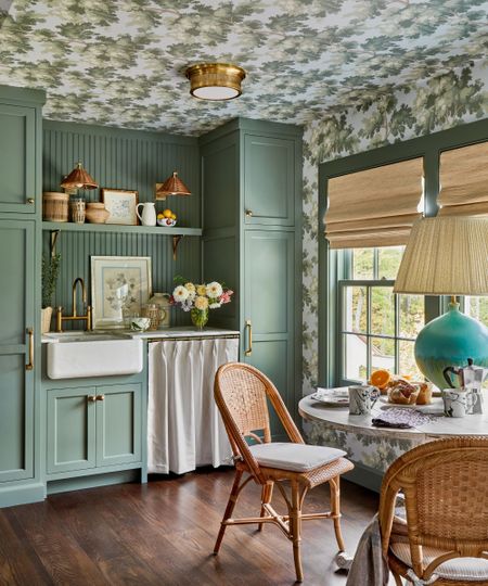 A kitchen with dark green cabinets, a patterned wallpapered ceiling, cozy rustic decor and a round table with two chairs.