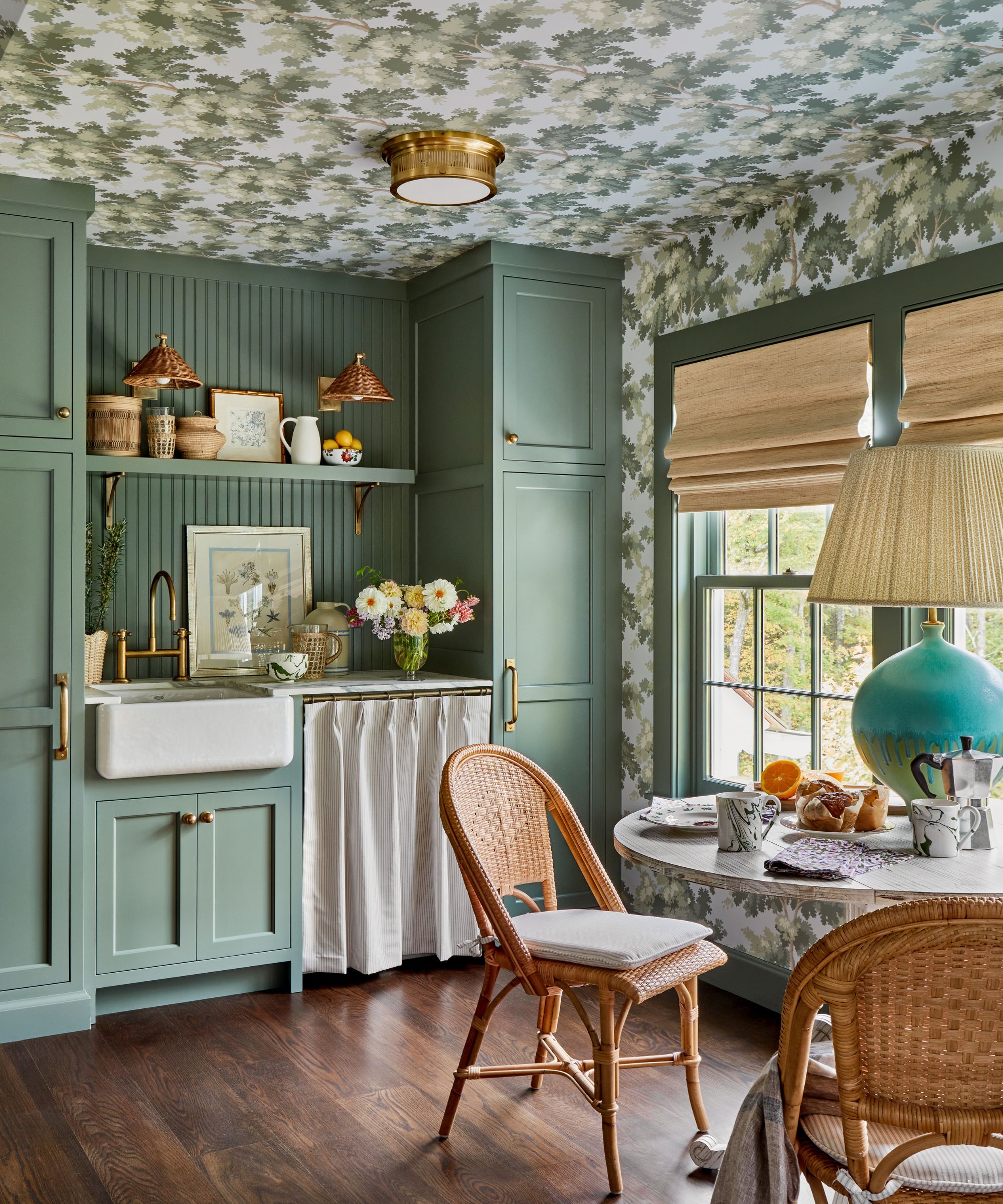 A kitchen with dark green cabinets, a patterned wallpapered ceiling, cozy rustic decor, and a round table with two chairs.
