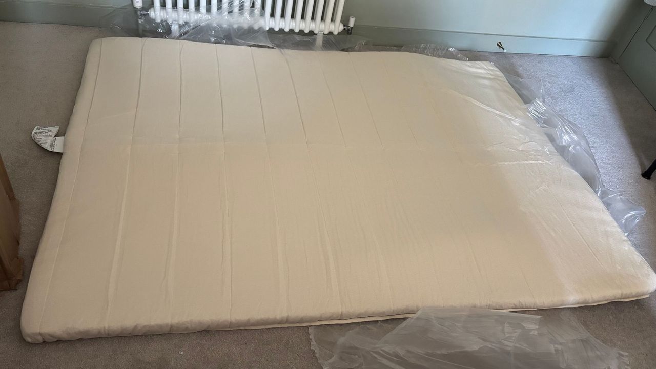 The freshly-unboxed Birch Plush Organic Mattress Topper still partially in its plastic wrapping and left on a carpeted floor to fully expand.