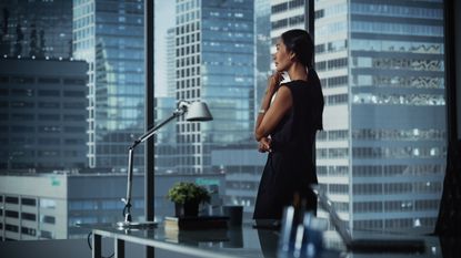 A businesswoman stands at the window of her office and looks out on a city skyline.