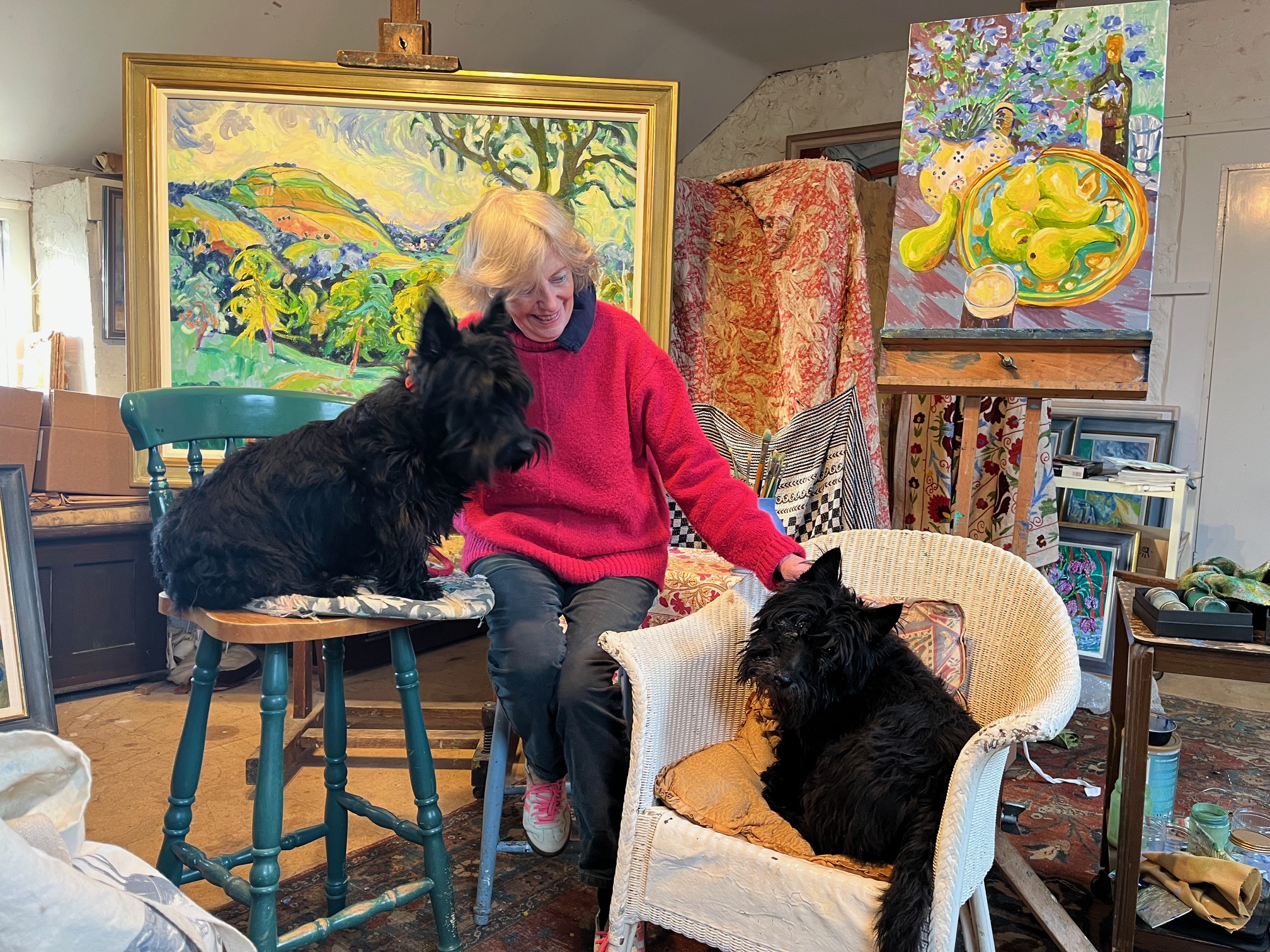Artist Josephine Trotter sits in her studio, smiling as she reaches out to her two Scottish terriers, Peebles and Presley &amp;mdash; one perched on a chair beside her, the other lounging in a wicker armchair &amp;mdash; with colourful framed paintings and an easel-filled workspace behind them.