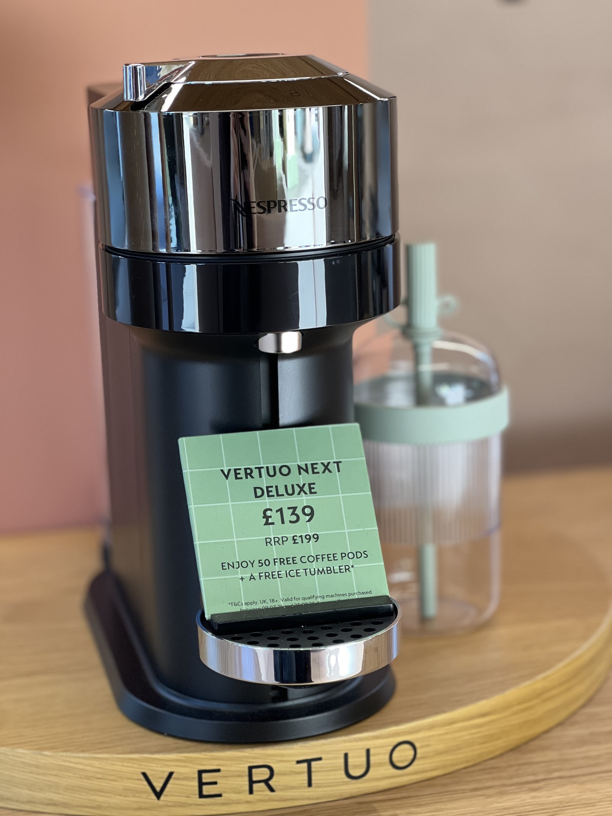 Image of a chrome and black coffee machine on a round, wooden base in a showroom. There is a plastic takeaway cup next to it.