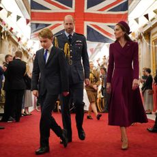 Prince George walking in front of Prince William in a military uniform and Prince Kate in a burgundy coat with a Union Jack behind them