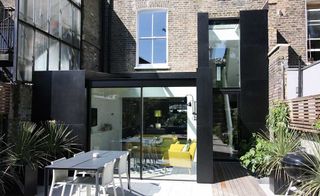 IQ glass contemporary black extension added onto a brick terraced house, leading out onto a white and wooden deck, with an outdoor dining table
