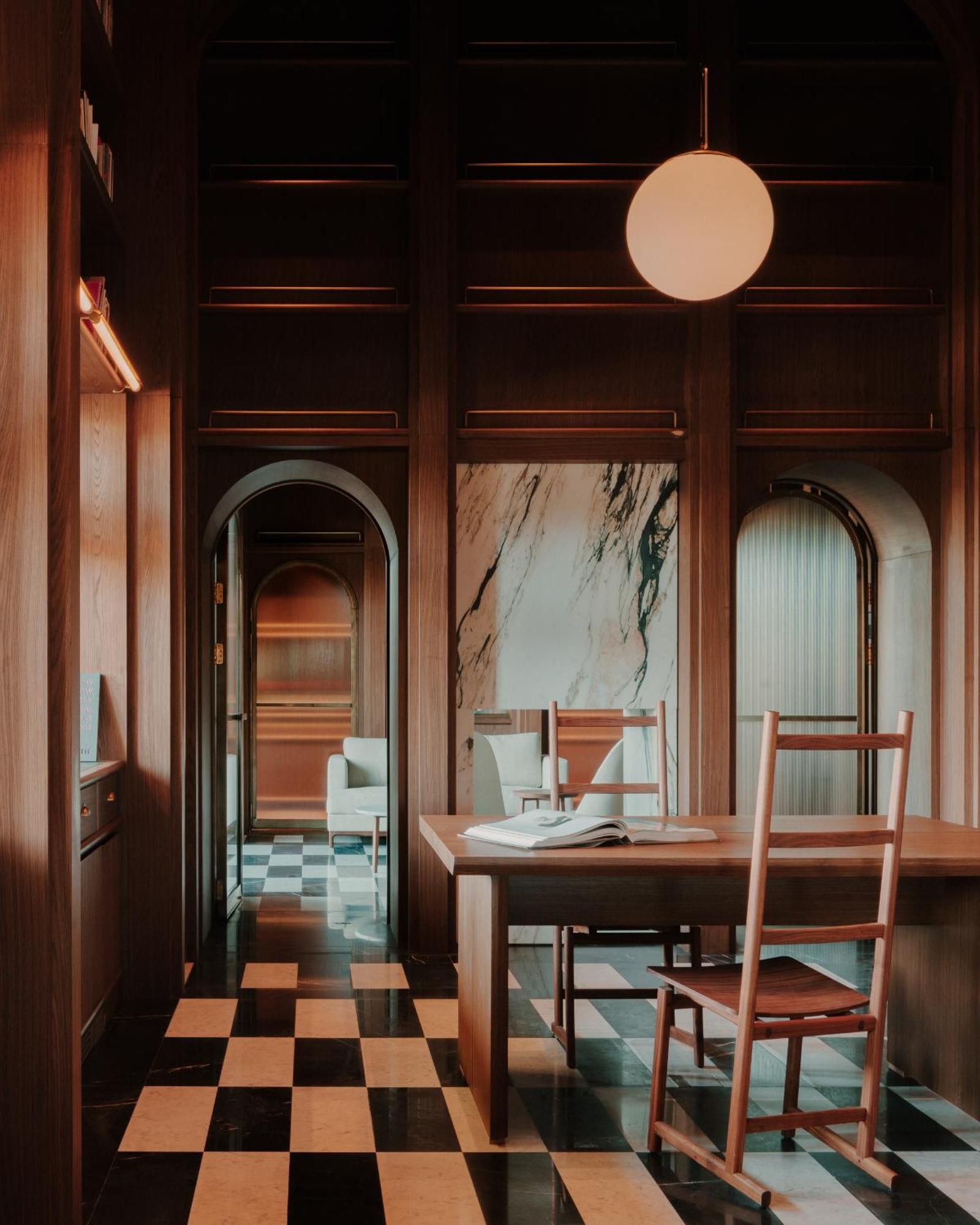 The brutalist-warm interiors of a luxury hotel sitting inside a former Soviet-era office feature checkered black and white, or wooden, floors, spherical soft lighting, and wooden furniture throughout.