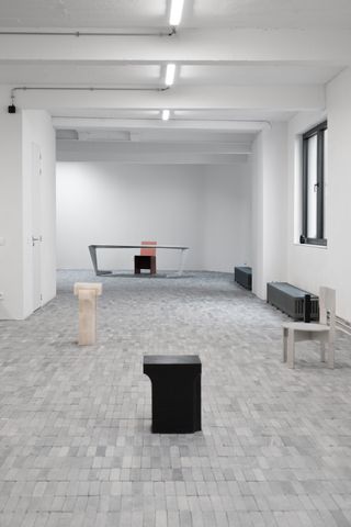 Alternative view of Studio Khachatryan's works on display against a white wall and stone tiled floor - works include dark, patinated bronze candleholders, aluminium desk and chair and onyx coffee table