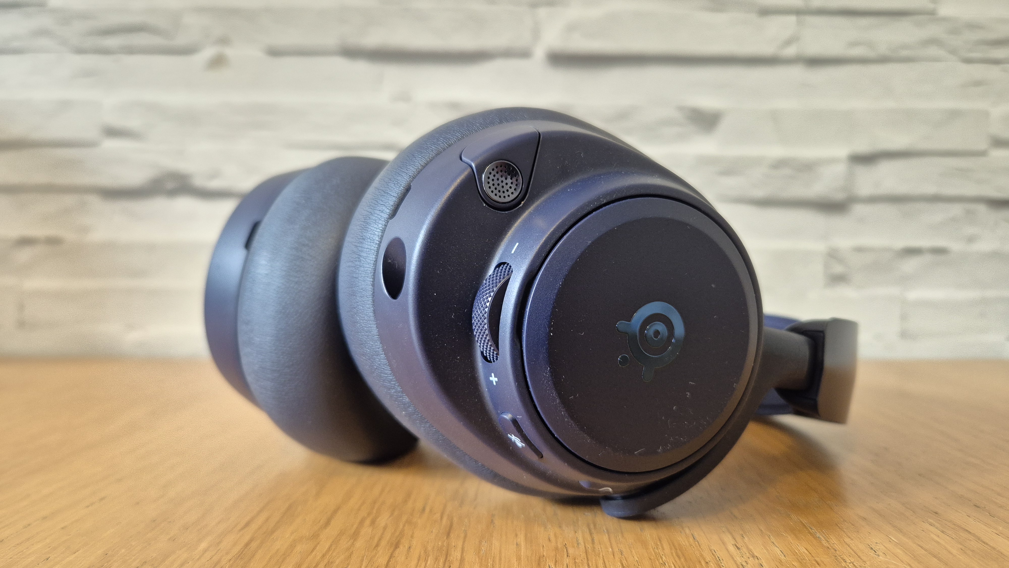 The SteelSeries Arctis Nova Pro Omni in front of a white brick background and on a wooden surface.