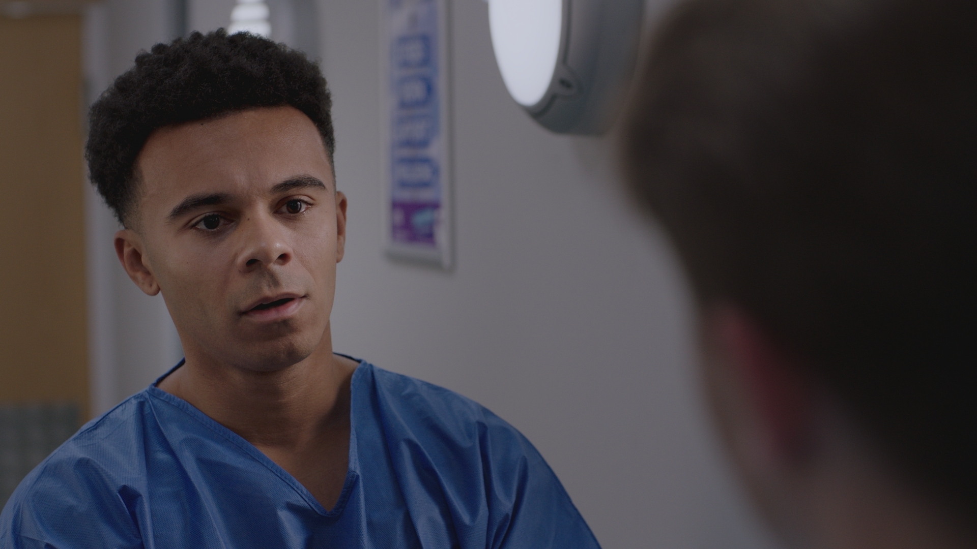 Teddy Gowan looking shaken wearing a set of blue scrubs and talking to someone off camera in Holby ED.