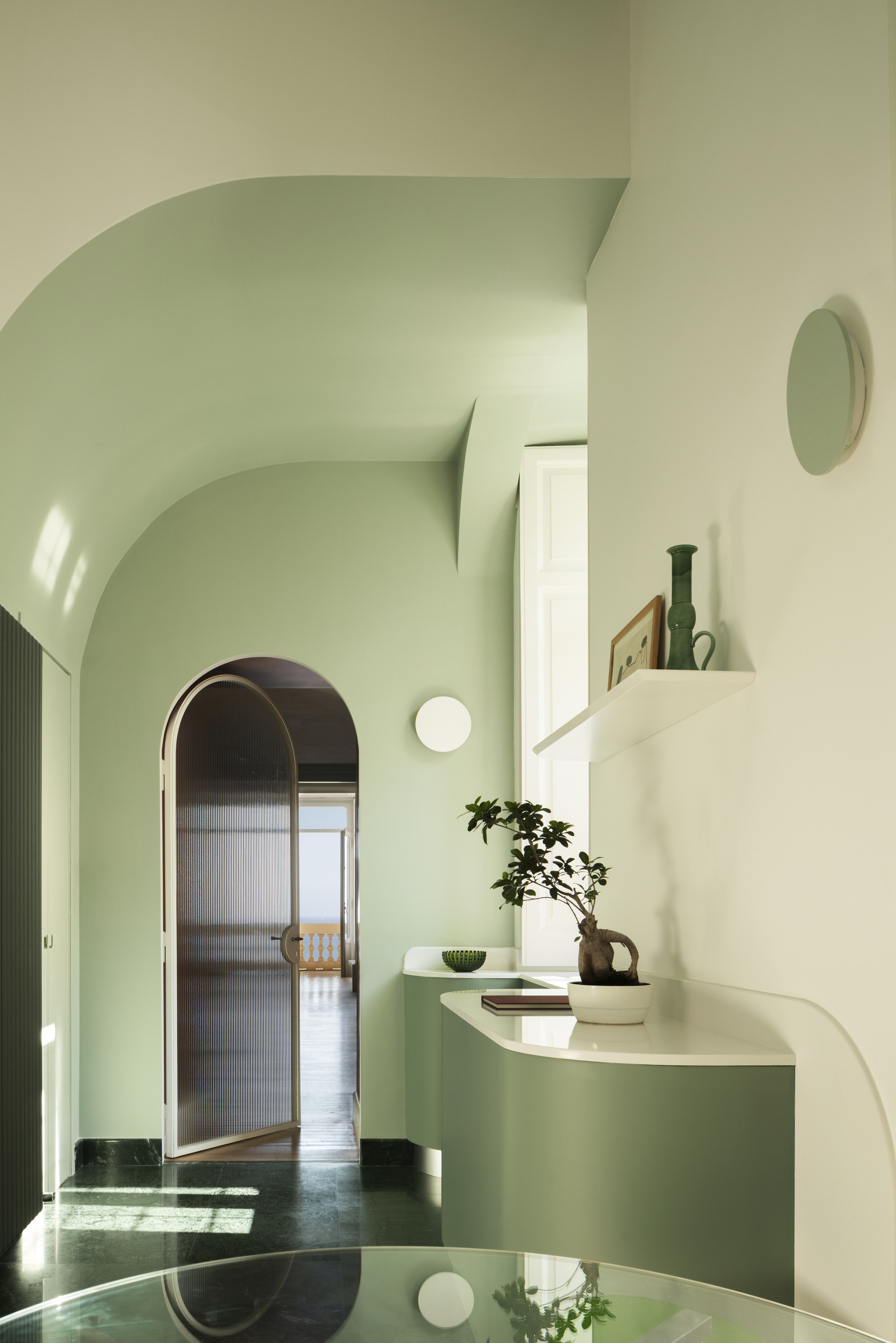 Image of a minty green hallway with curved ceiling and curved cabinetry. There is a white bowl and a plant in a vase on the counter.