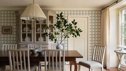 Sun-drenched dining room featuring blue-and-white rose-motif wallpaper, airy white dining chairs, a pleated pendant light, and a large potted plant