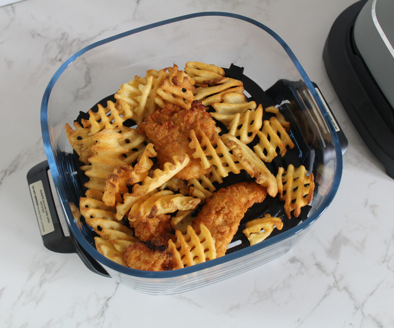 A glass air fryer dish viewed from above, filled with waffle fries and breaded chicken.
