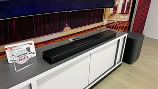 En svart JBL Bar 1300X soundbar ligger placerad på en TV-bänk under en TV på CES 2023.