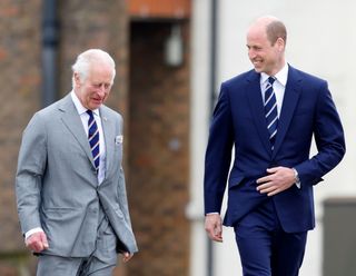 Prince William wearing a blue suit and The King in a gray suit walking next to each other outside and smiling