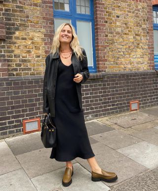 British influencer Lucy Williams smiles as she poses on a London sidewalk wearing multi-strand pearl necklaces, a black leather jacket, a black slip dress, a black bag and olive green chunky loafers