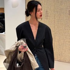style influencer Mimi Nguyen poses with a low bun, red lipstick, black collarless cinched-waist blazer, and jeans with a trench coat and brown woven leather bag