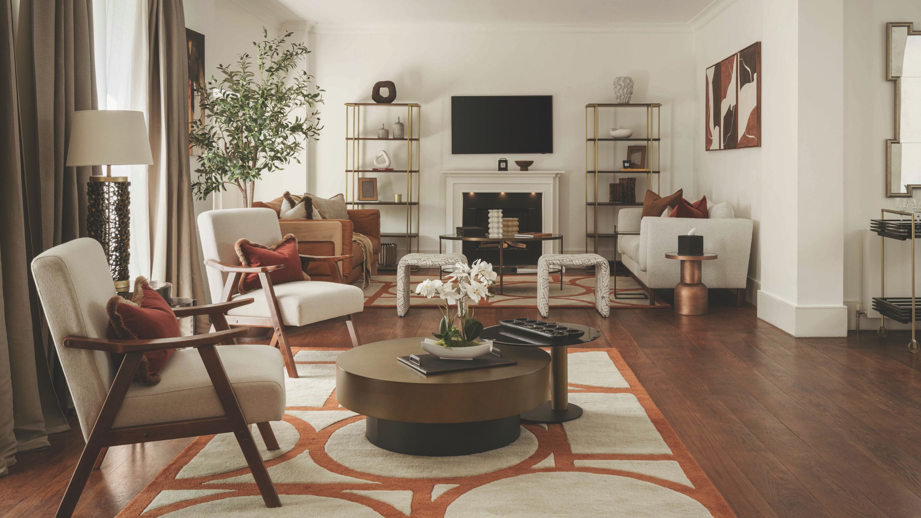 An elegant mid-century modern living room with deep green walls and terracotta-patterned rugs. Two beige armchairs and a copper-hued coffee table are positioned in the foreground. A white fireplace with open gold shelving is at the back.
