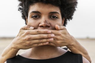 A Black woman facing and staring the camera. There are hands over her mouth and on her nose is some hyperpigmentation