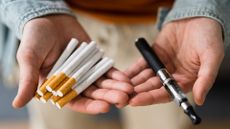 Close up of a pair of hands holding smoking parafernalia. In the left hand is a vape pen and in the right hand there is a handful of cigarettes.