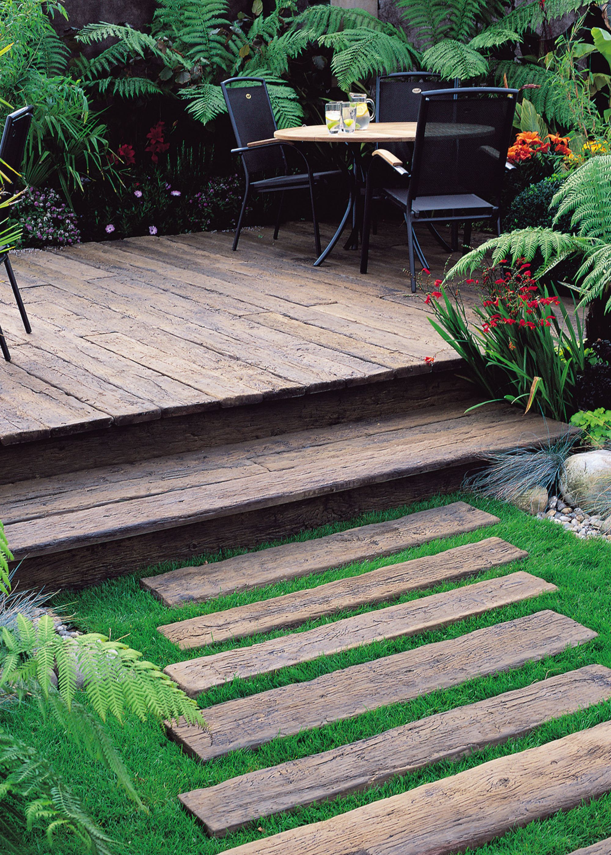 A garden with wooden steps and wooden decking. There are plants and trees all around as well as a view of a patio dining table ad three chairs around it.