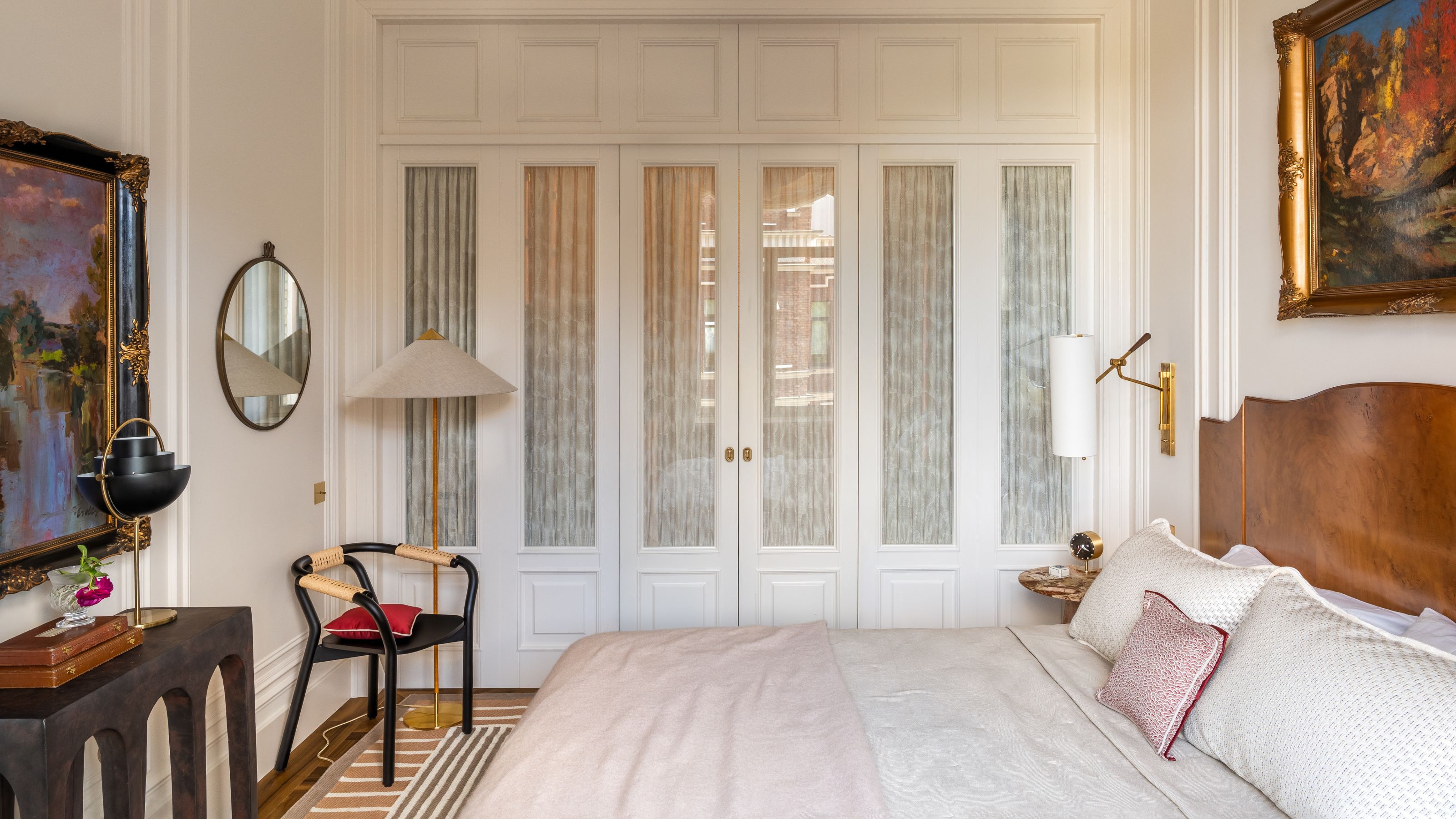 Image of a formal, white bedroom with french doors leading to a closet and a blush and beige bed that has a wooden head board. There is classic art on the walls, a console table, and vintage lamps and mirrors.