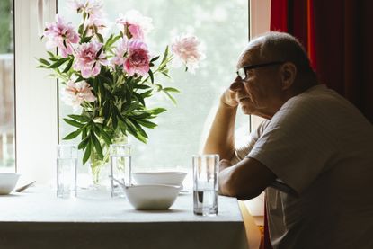 Portrait of a serious, sad elderly man with glasses. He sits at a dining room table near a window. There are pink peonies at the dinner table. 