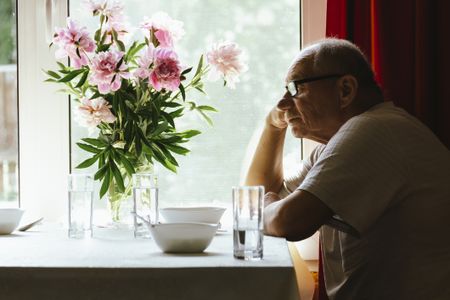 Portrait of a serious, sad elderly man with glasses. He sits at a dining room table near a window. There are pink peonies at the dinner table. 