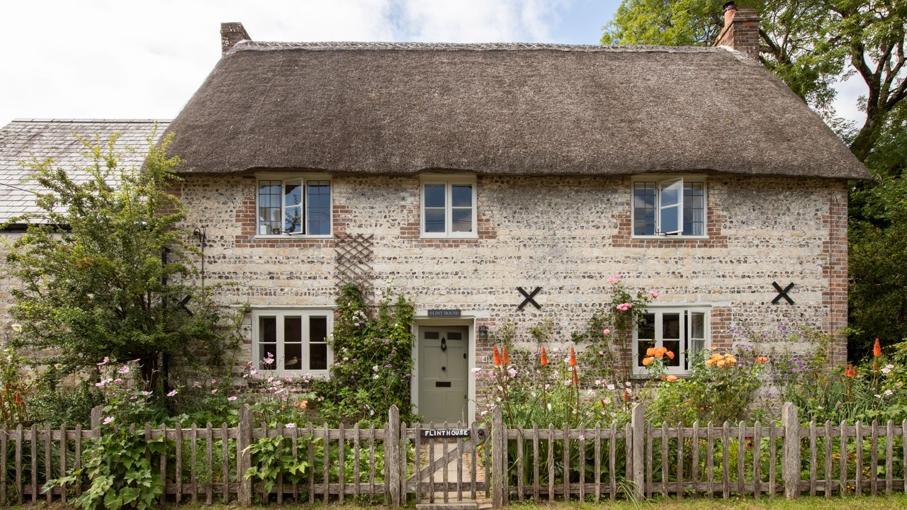 exterior of a flint stone thatched cottage with flowers in front garden and picket fence