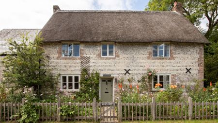 exterior of a flint stone thatched cottage with flowers in front garden and picket fence