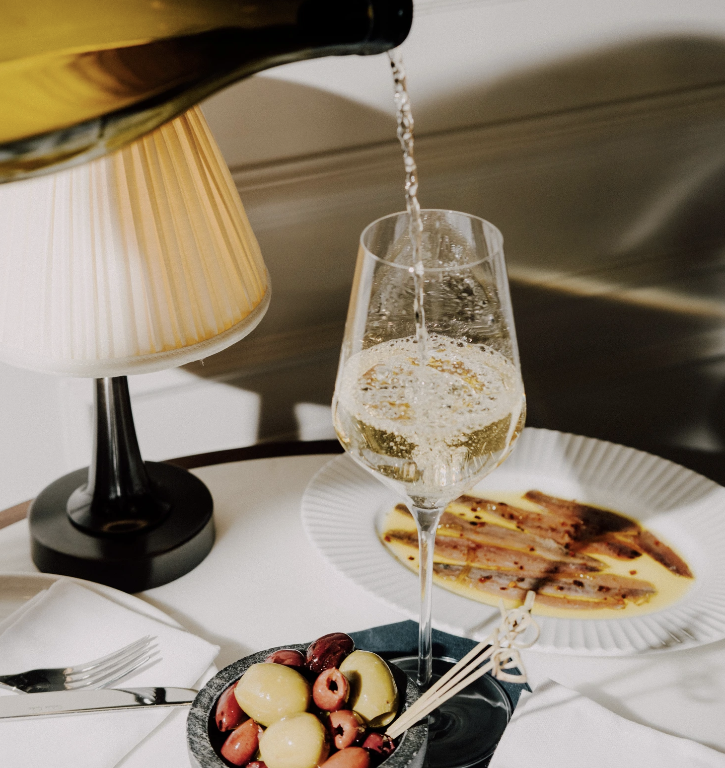 an image of white wine being poured into a wine glass, alongside two plates of food on the same table