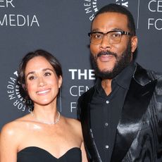 Meghan Markle wears a black strapless dress while posing with Tyler Perry who is dressed in a black suit and shirt