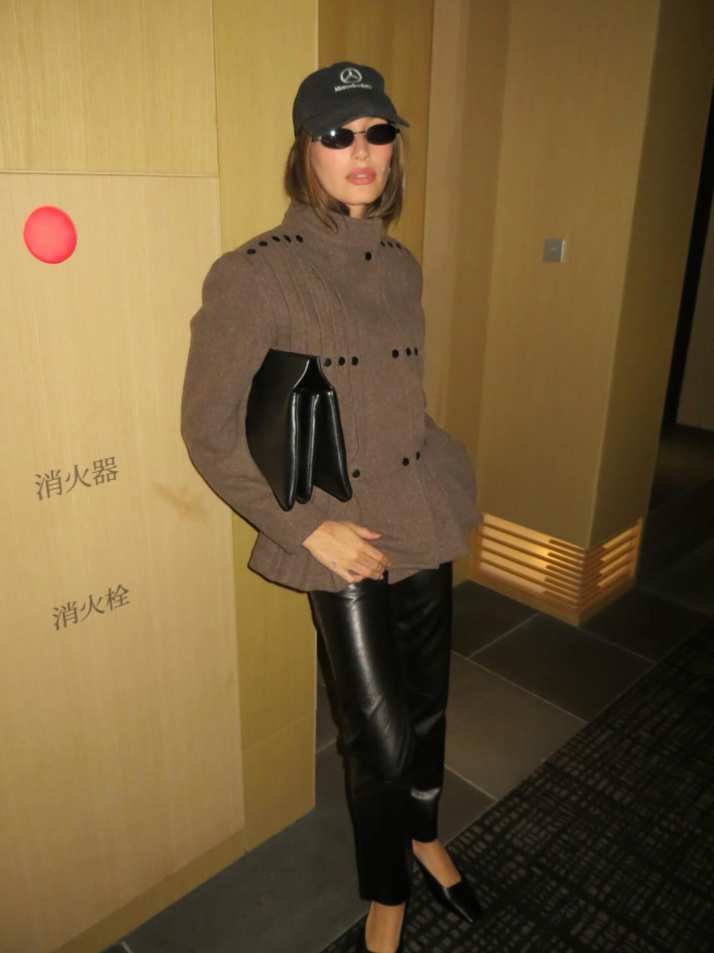 Hailey Bieber in Tokyo wearing a Mercedes baseball cap, brown jacket, and black leather pants.
