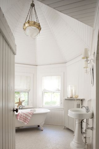 white bathroom with vaulted ceiling, shiplap walls and ceiling, white roll top tub, curvy basin, small hexagon floor tiles, wall lights, radiator, pendant light