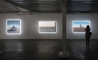 Art pieces on canvases are placed throughout the room, with a light on them. A woman is looking at the art piece to the right. The rest of the space is dark.