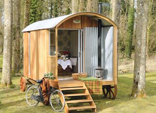 a shepherd's hut in a wooded area full of trees, with steps leading up to it and a bicycle out front, and a dining table and chairs set up inside