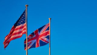 The UK and US flag flying on flagpoles, with the US flag to the left of the UK flag, shot against a blue sky. Decorative: the warm light on the flags suggests it is sunrise or sunset.