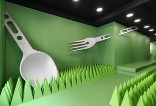 A large spoon resting against a wall. A large fork attached to the wall. The walls are floor are green. Virgil Abloh cutlery by Alessi installation at Galleria Manzoni