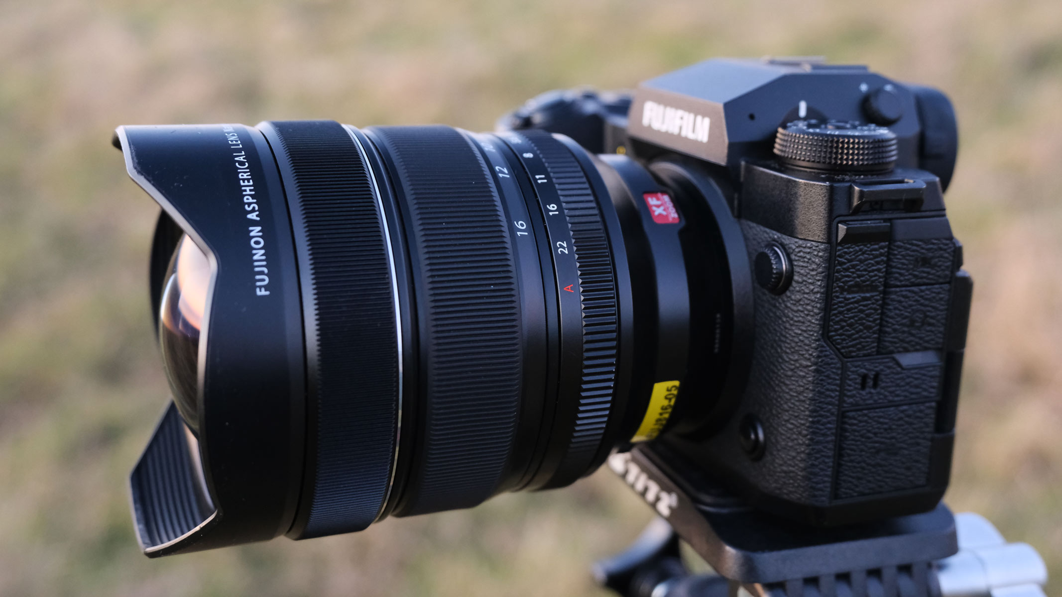 A side view of the Fujifilm X-H2 with the Fujifilm XF 8-16mm f/2.8 lens attached and a field in the background